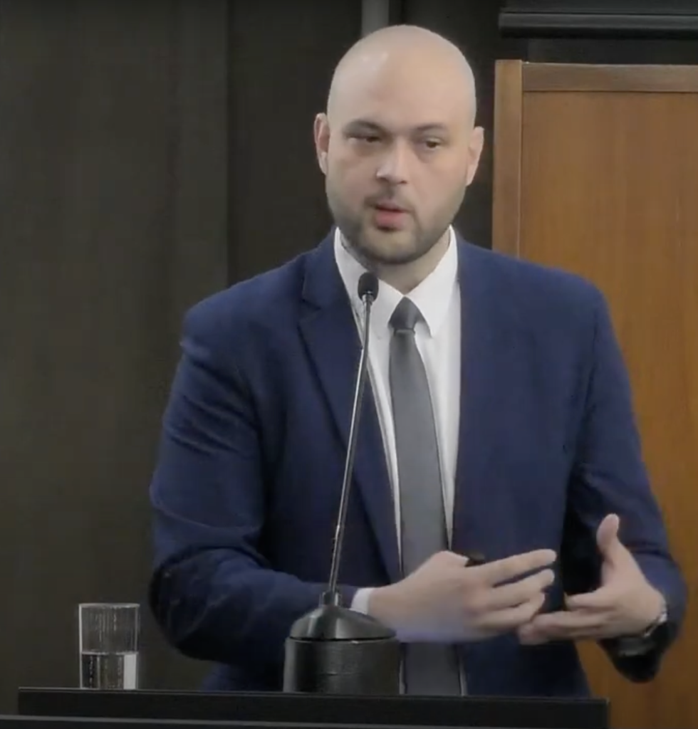 Jonas Gomes, vestido com um terno azul escuro, camisa branca e gravata cinza, apresenta-se em um púlpito, gesticulando enquanto fala. Ele está em um ambiente formal, com um copo de água à sua esquerda sobre o púlpito e móveis de madeira ao fundo. A imagem transmite um momento de sua palestra, destacando sua comunicação clara e envolvente.